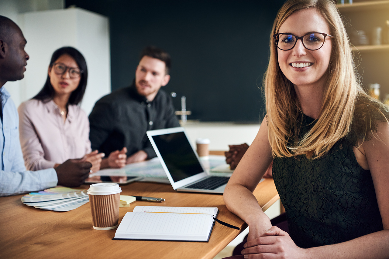 Smiling young businesswoman sitting at a boardroom table during a meeting with colleagues in the background