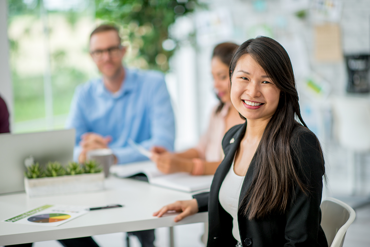 A group of businesspeople are indoors in an office. A woman is smiling at the camera.