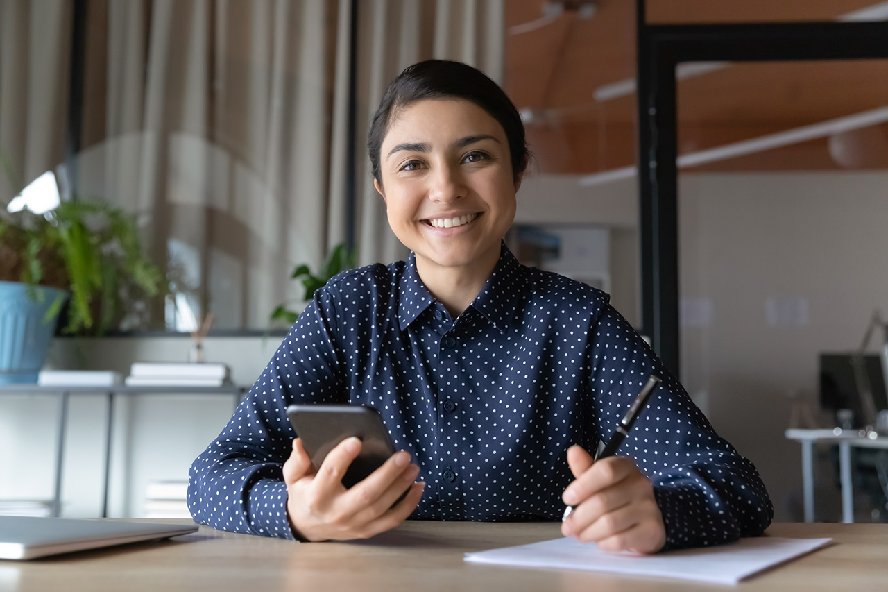 Portrait of smiling indian female student sit at desk make notes watching webinar on smartphone. Happy young mixed race ethnicity woman study take online course or training using cellphone gadget.