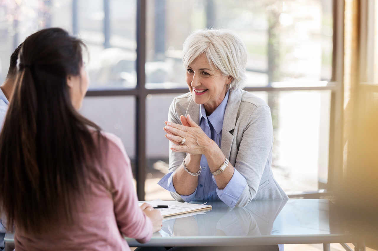 A smiling mature female counselor sits at a table across from an unrecognizable couple.  She puts her hands together as she listens to the woman.
